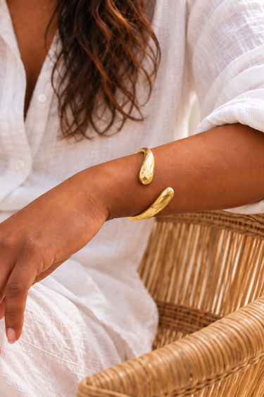 Close-up of a Nyoka Hammered brass bangle on a wrist resting on a wicker chair.