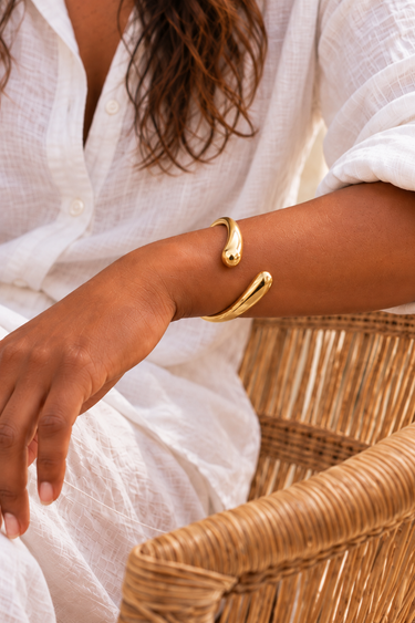 Close-up of a Nyoka Smooth brass bangle on a wrist resting on a wicker chair.